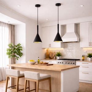 Puede incluir: Dos lámparas colgantes negras en forma de cono con interiores dorados cuelgan sobre una isla de cocina. La cocina cuenta con gabinetes blancos, una isla de madera con dos taburetes beige y una estufa de acero inoxidable. Se ve una planta verde y una ventana con una cortina transparente.