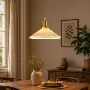 May include: A retro pendant light with a ribbed glass shade and brass fittings hangs above a wooden dining table. The light is suspended from a woven cord. The room is decorated in neutral tones with a vase of flowers and a framed botanical print.