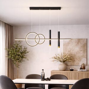 Può includere: Un moderno lampadario nero e oro con tre luci a sospensione. Il design comprende due cerchi interconnessi, due luci cilindriche e una luce lineare. Il lampadario è sospeso sopra un tavolo da pranzo.