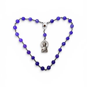 May include: A rosary featuring faceted deep purple beads strung on a dark metal chain. The piece includes a central silver-toned metal connector and a circular pendant depicting a figure in relief, arranged in a heart-shaped formation.