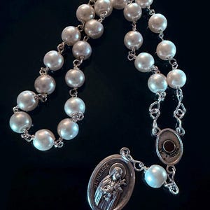 Può includere: Un rosario con perle bianche madreperlace collegate da maglie in metallo argentato. Il rosario include un elemento centrale ovale in metallo argentato e un ciondolo che raffigura una figura con il testo inciso ST. THERESE sul davanti.