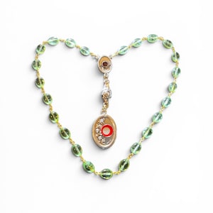 Op de afbeelding: Een rozenkrans met doorschijnende lichtgroene ovale kralen verbonden door goudkleurig draad. Het middelpunt is een zilverkleurige ovale medaille met een rood-witte cirkelvormige inleg, een bloemengravure en een zilverkleurig roosaccent erboven.