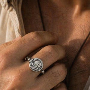 Könnte beinhalten: Ein silberner Ring mit einem kreisförmigen Anhänger mit der Aufschrift "PADRE" und dem Namen "PIO". Der Anhänger zeigt ein detailliertes Porträt. Der Ring wird an einem Finger getragen.