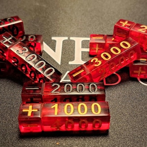 May include: Translucent red game chips with gold and silver numbering, displaying values such as 1000, 2000, 3000, and 4000, alongside plus and minus symbols. The chips are scattered on a dark surface.