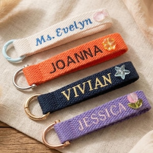 Pode incluir: Quatro chaveiros personalizados em creme, laranja, azul marinho e roxo. Cada chaveiro apresenta um nome bordado em uma cor contrastante: Sra. Evelyn, Joanna, Vivian e Jessica. Os enfeites decorativos incluem uma estrela, uma flor e uma gema.