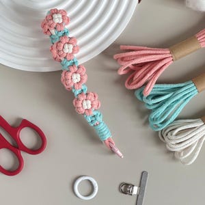 May include: A handmade macrame flower bookmark with pink and white flowers and turquoise accents. The bookmark is next to bundles of pink, turquoise, and white macrame cord, red scissors, a white ring, and a silver clip.