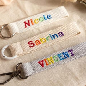 May include: Three fabric keychains with embroidered names. Two are off-white with the names Nicole and Sabrina in rainbow-colored thread, featuring silver-toned rings. One is light gray with the name VINCENT in rainbow thread and a dark metal clasp.