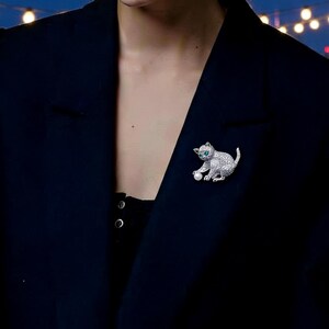 Può includere: Un fermaglio a forma di gatto argentato con una piccola perla e occhi verdi è appuntato su un blazer blu scuro. Il fermaglio è incastonato con piccole pietre scintillanti. Lo sfondo presenta un paesaggio urbano sfocato e luci a catena.