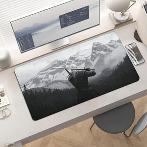 May include: A black and white rectangular desk mat featuring a warrior with a sword in a mountain landscape. The mat is designed to protect the desk surface and provide a smooth surface for a mouse.