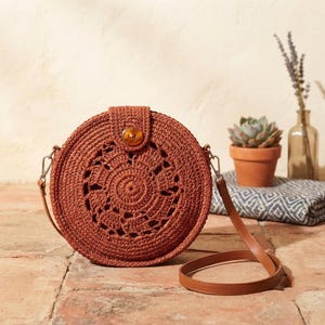 May include: A round, rust-colored crochet crossbody bag featuring a central circular pattern with openwork details. The bag has a flap closure secured by a single brown button and includes a thin, smooth brown leather-like shoulder strap.