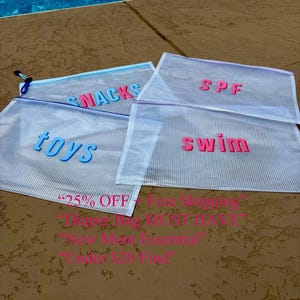 May include: Clear mesh zippered pouches labeled "SNACKS", "TOYS", "SPF", and "swim" in blue and pink lettering. The pouches are arranged on a brown surface near a pool. A blue and black zipper pull is visible.