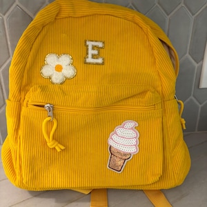 Puede incluir: Una mochila de pana de color amarillo brillante con un bolsillo frontal con cremallera, correas ajustables y tres parches decorativos: una flor blanca con centro amarillo, la letra E en una fuente con borde dorado y un helado de lentejuelas.