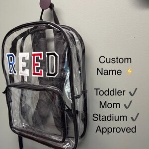 May include: A clear plastic backpack with black trim and black straps, featuring the name REED in large varsity-style letters with blue, white, red, and black colors. Text on the right reads Custom Name, Toddler, Mom, Stadium, and Approved.