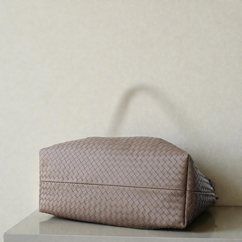 May include: A taupe handbag with a woven design. The bag is rectangular with a flat base and a handle. The bag is placed on a light-coloured surface.