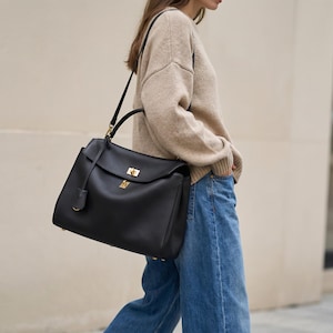 Könnte beinhalten: Eine schwarze Lederhandtasche mit strukturiertem Design. Die Tasche hat einen oberen Griff, einen Schulterriemen und goldfarbene Hardware. Ein kleines, passendes Lederetikett hängt vorne. Die Tasche wird von einer Person getragen, die einen beigen Pullover und blaue Jeans trägt.
