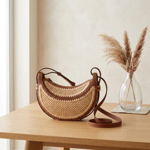 May include: A crescent-shaped handbag with a woven straw center and brown leather trim. The bag has a long, adjustable strap and a zipper closure. It sits on a wooden table next to a vase of dried flowers, showcasing a natural, bohemian aesthetic.