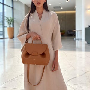 K&ouml;nnte beinhalten: Eine braune Lederhandtasche mit geschwungenem &Uuml;berschlag und Tragegriff. Die Tasche hat einen Schulterriemen und goldfarbene Beschl&auml;ge. Die Tasche wird von einer Person getragen, die ein beiges Kleid tr&auml;gt.