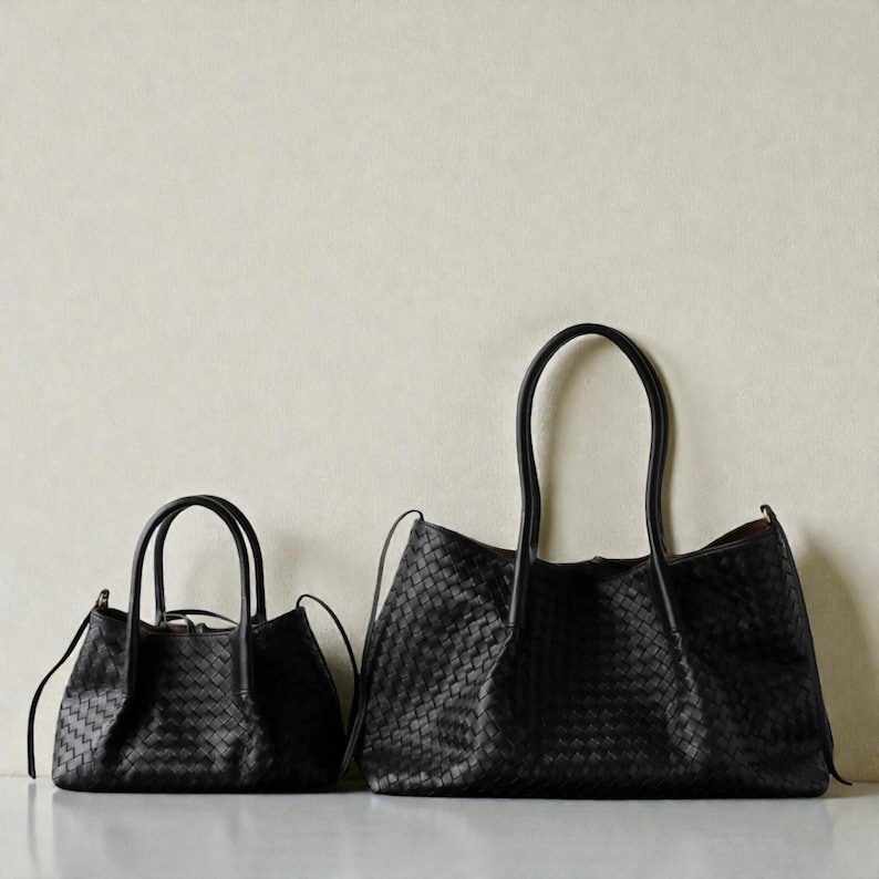 May include: Two black leather handbags with a woven design. The smaller bag has a top handle and a shoulder strap. The larger bag has long handles and a wider opening. Both bags are made of dark, matte leather.