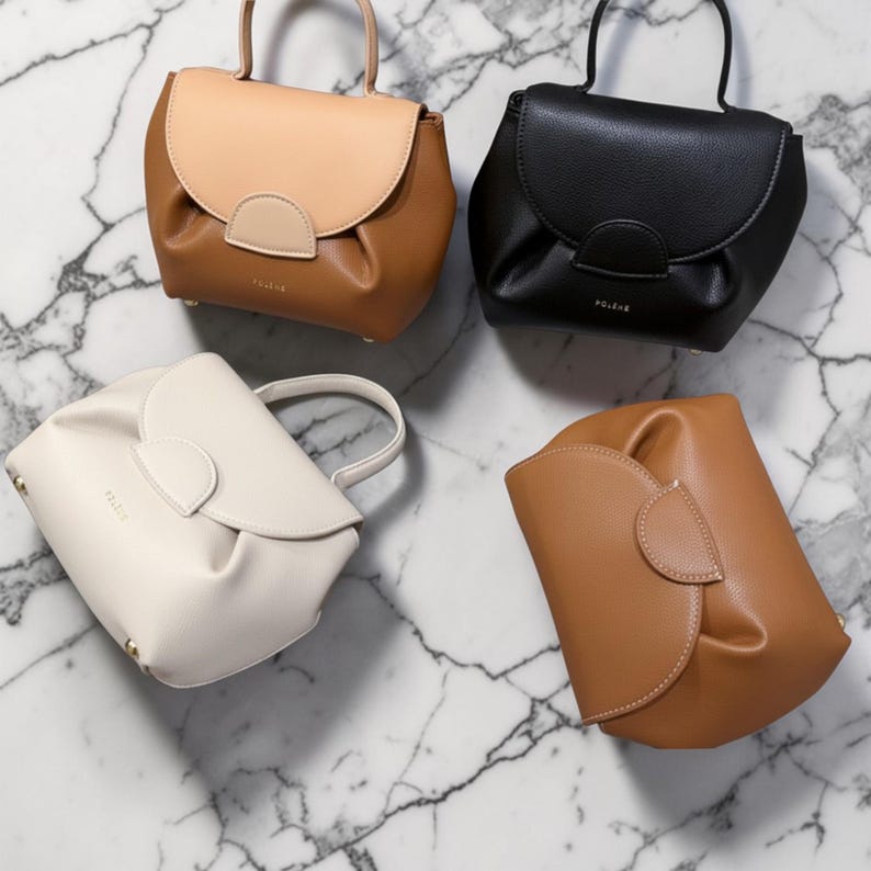 K&ouml;nnte beinhalten: Vier kleine Lederhandtaschen in verschiedenen Farben: Beige, Schwarz, Wei&szlig; und Braun. Jede Tasche hat einen geschwungenen Klappenverschluss und einen oberen Griff. Die Taschen sind auf einer Marmoroberfl&auml;che angeordnet.