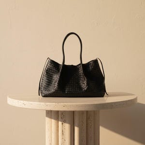 May include: A black woven leather tote bag with long handles rests on a round, light-coloured stone table. The bag has a structured shape and a spacious interior. The background is a neutral beige colour, providing a clean and elegant aesthetic.