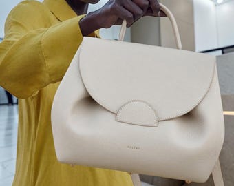 May include: Cream-colored handbag with a rounded flap and top handle. The bag has a structured design with a subtle embossed logo. The bag is held by a person wearing a yellow shirt. The bag has a shoulder strap.