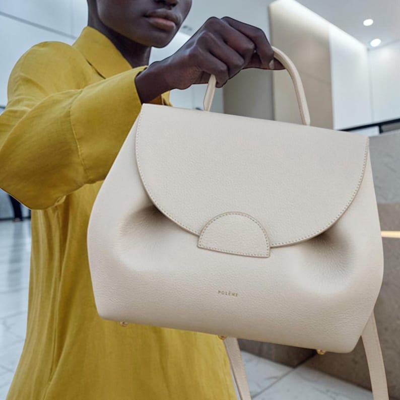 K&ouml;nnte beinhalten: Cremefarbene Handtasche mit abgerundeter Klappe und oberem Griff. Die Tasche hat ein strukturiertes Design mit einem dezent gepr&auml;gten Logo. Die Tasche wird von einer Person gehalten, die ein gelbes Hemd tr&auml;gt. Die Tasche hat einen Schultergurt.