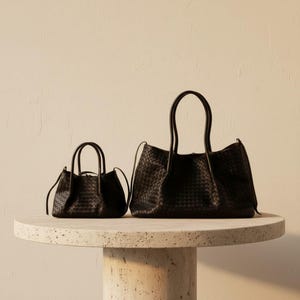 May include: Two black woven leather handbags of different sizes are displayed on a round, light-coloured table. The larger bag has two handles and a wider opening, while the smaller bag has a more compact design with a single handle.