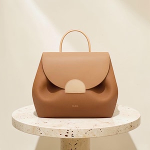K&ouml;nnte beinhalten: Braune Lederhandtasche mit runder Form und oberem Griff. Die Tasche hat einen Klappenverschluss mit hellbraunem Akzent und den Markennamen "POLENE" auf der Vorderseite. Die Tasche steht auf einem Marmortisch.