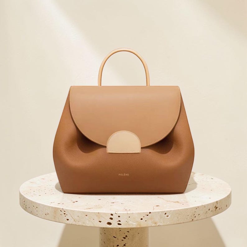 K&ouml;nnte beinhalten: Braune Lederhandtasche mit runder Form und oberem Griff. Die Tasche hat einen Klappenverschluss mit hellbraunem Akzent und den Markennamen "POLENE" auf der Vorderseite. Die Tasche steht auf einem Marmortisch.