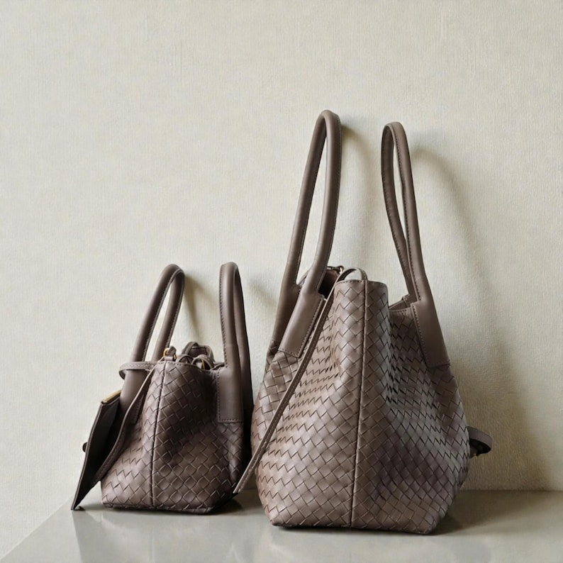 May include: Two taupe-coloured handbags with woven leather exteriors. The bags have long, curved handles and a structured design. One bag is larger with a taller handle, while the other is smaller with a shorter handle. Both bags are sitting on a reflective surface.