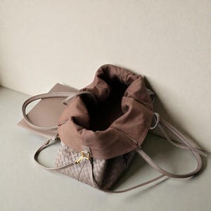 May include: A taupe-coloured woven leather tote bag with a brown interior. The bag has long, thin handles and a gold-coloured clasp. A matching clutch is visible in the background.