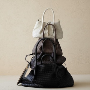 May include: Four woven leather handbags in a stack. The top bag is white, followed by brown, a darker brown, and a black bag at the bottom. Each bag has a handle. The bags are in a neutral setting.