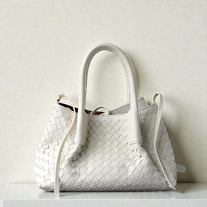 May include: A white handbag with a woven pattern. The bag features two curved handles and a long strap. The bag is placed on a white surface.