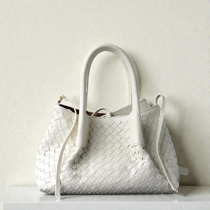 May include: A white handbag with a woven pattern. The bag features two curved handles and a long strap. The bag is placed on a white surface.