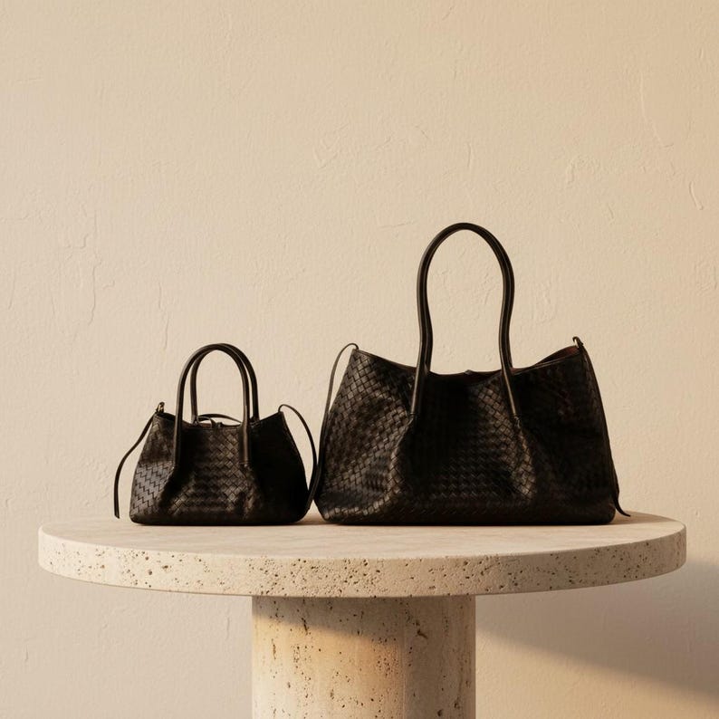May include: Two black woven leather handbags of different sizes are displayed on a round, light-coloured stone table. The larger bag has two handles and a wider opening, while the smaller bag has a more compact design with a top handle.