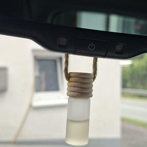 Könnte beinhalten: Eine kleine, zylindrische Flasche aus Milchglas, gefüllt mit einer blassen Flüssigkeit, die an einer braunen Kordel an einem schwarzen Rückspiegel eines Fahrzeugs hängt. Die Flasche hat einen gerippten, hellen Holzdeckel zur Befestigung.