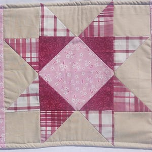 May include: A quilted placemat featuring a central star design in shades of pink and cream. The star is composed of various patterned fabrics, including floral and plaid prints. The edges are bordered with a floral pattern.