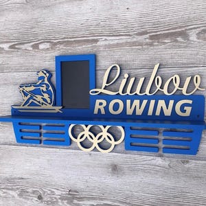 Puede incluir: Estante azul de madera con temática de remo con el nombre "Liubov" y la palabra "ROWING" en una fuente decorativa. Cuenta con un recorte de un remero, un pequeño marco de fotos y los anillos olímpicos. El estante está montado sobre un fondo de madera.