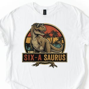 May include: White t-shirt featuring a retro design with a dinosaur wearing sunglasses. The design includes a circular background with palm trees and the text "SIX-A SAURUS" in a rectangular banner.