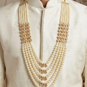 Peut inclure: Un collier de perles crème à plusieurs rangs avec des accents dorés et des pierres précieuses transparentes. Le collier est porté sur un vêtement crème. Il comporte plusieurs rangs de perles, avec des éléments décoratifs en or et en pierres précieuses.