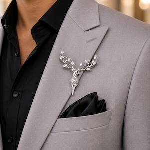 Peut inclure: Une broche en forme de cerf argenté incrustée de cristaux transparents, avec une tête détaillée et des bois ramifiés ornés de pierres taille marquise. La broche est épinglée sur le revers d'un blazer gris clair sur une chemise noire.