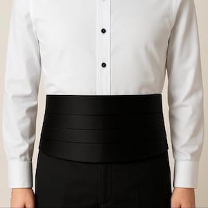 May include: A black cummerbund with four pleats is worn over black trousers. The cummerbund is paired with a white dress shirt with black buttons. The formal wear is suitable for a tuxedo or other formal occasions.