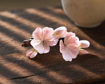 Puede incluir: Una pinza para el cabello con dos delicadas flores de color rosa pálido con vetas rojas sutiles en los pétalos. El accesorio floral está sujeto a una base rectangular de color marrón oscuro, mostrando pétalos suaves y redondeados y capullos.