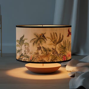 May include: A table lamp with a cylindrical lampshade featuring a jungle scene with animals and lush foliage. The base is a light orange color, and the shade has a black trim. The lamp is illuminated, casting a warm glow.