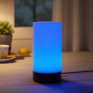 Könnte beinhalten: Zylindrische Tischlampe mit schwarzem Sockel und blauem Licht. Die Lampe steht auf einer Holzoberfläche, im Hintergrund eine Tasse, Untertasse und Zitrone. Ein schwarzes Kabel geht von der Lampe ab.