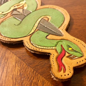 Peut inclure: Un écusson en cuir fait à la main représentant un serpent vert enroulé autour d'un poignard. Le serpent a une langue rouge et des yeux jaunes. L'écusson est de couleur beige avec des accents dorés et une bordure cousue.
