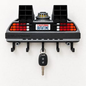 Puede incluir: Un soporte de pared para llaves diseñado como la parte trasera de un coche DeLorean, con rejillas negras, luces traseras rojas y blancas, y una matrícula de California que dice OUTATIME. La unidad tiene cinco ganchos negros con una llave colgada.