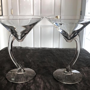 Puede incluir: Dos copas de martini de cristal transparente con un diseño de tallo curvo único. Las copas tienen una forma de cuenco triangular clásico y se asientan sobre una base redonda. Los tallos se curvan con gracia, añadiendo un toque moderno a la cristalería.