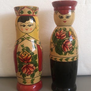 May include: Two hand-painted wooden nesting dolls. One doll is yellow with a red base and red accents, featuring a floral design. The other doll is black at the base, with a red hat and floral design. Both dolls have painted faces.