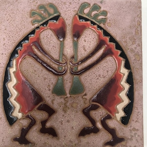 May include: A square ceramic tile featuring a textured, dusty pink background with a symmetrical design of two stylised figures playing flutes. The figures are rendered in shades of dark red, black, green, and cream, with arched bodies and feathered headdresses.